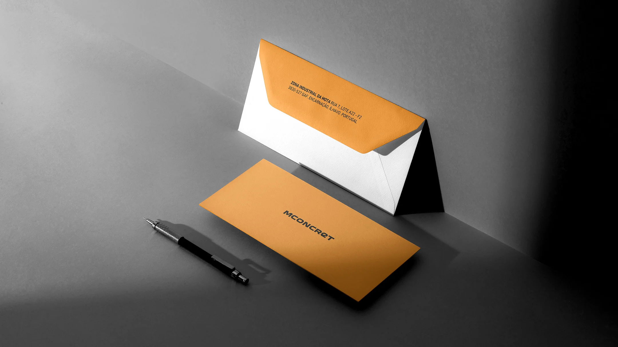 Projeto Mconcret da Nhdesign: Envelopes corporativos da Mconcret em laranja e branco, acompanhados de caneta.